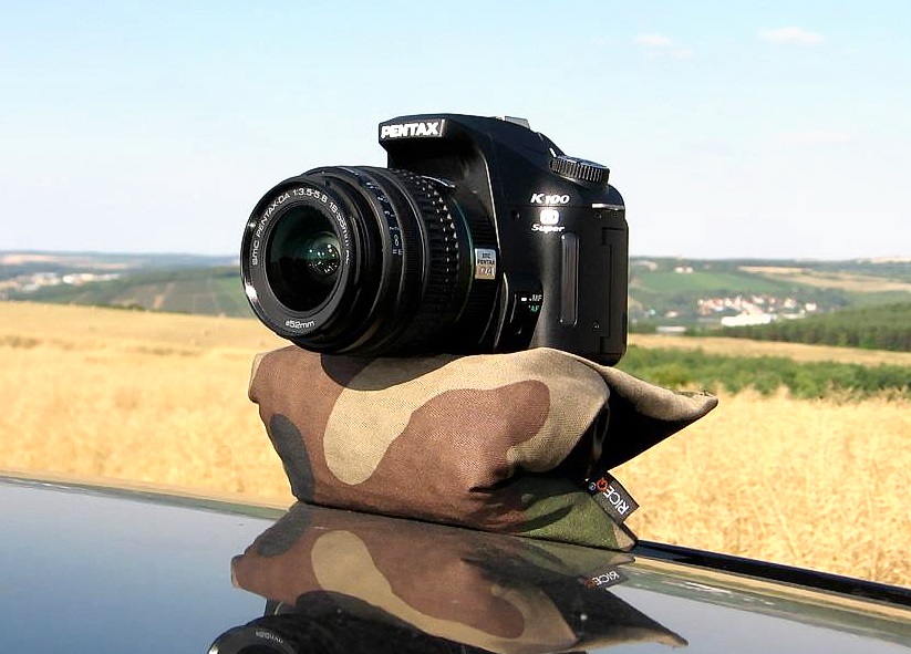 Kamera auf dem RiceQ Bohnensack Outdoor Oliv im Tarnmuster auf einem Autodach, Feld im Hintergrund